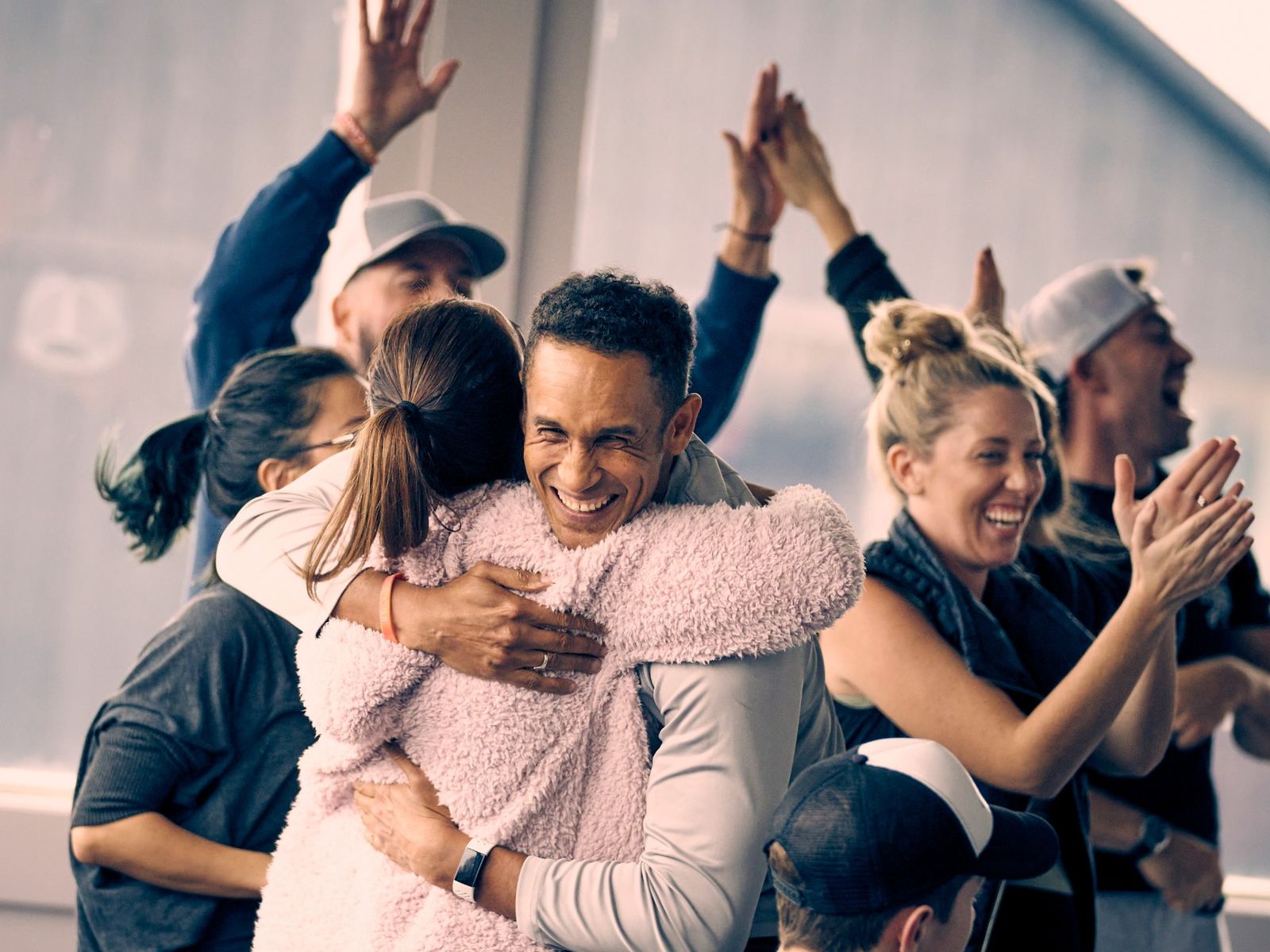 Two people hugging and laughing while a group behind them cheers with their hands in the air