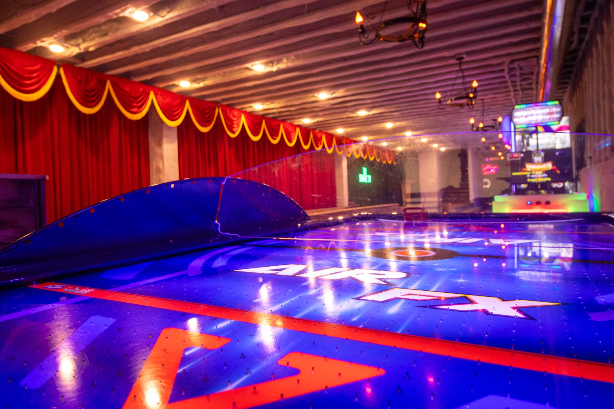 An illuminated air hockey table in the basement, red curtains and arcade cabinets behind it