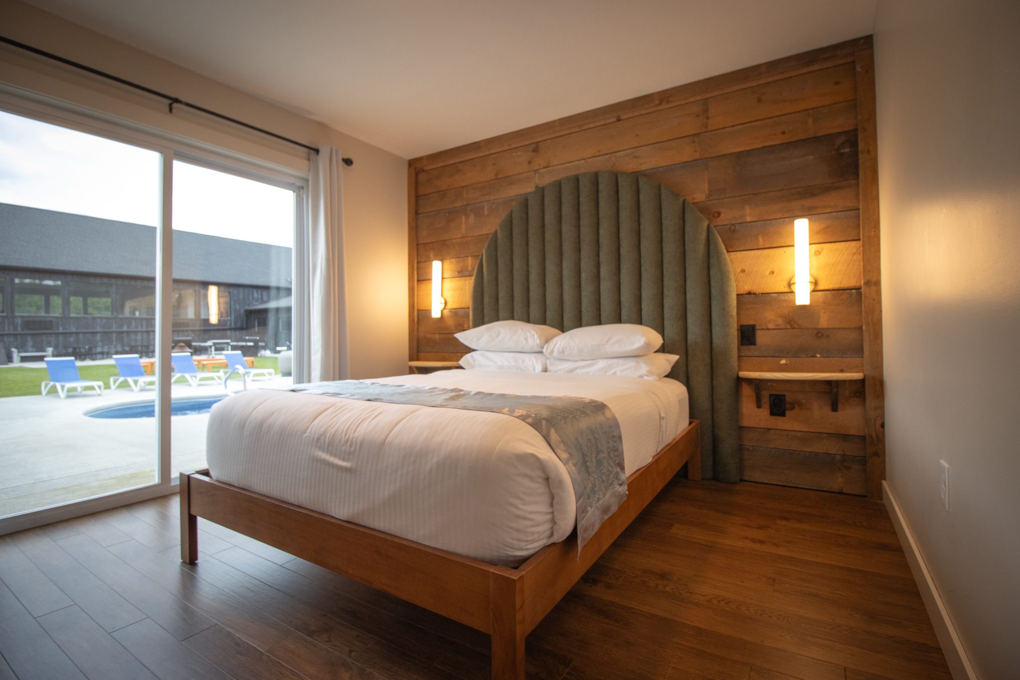 A queen bedroom with an arched upholstered headboard, a timber wall and a sliding door to the pool deck