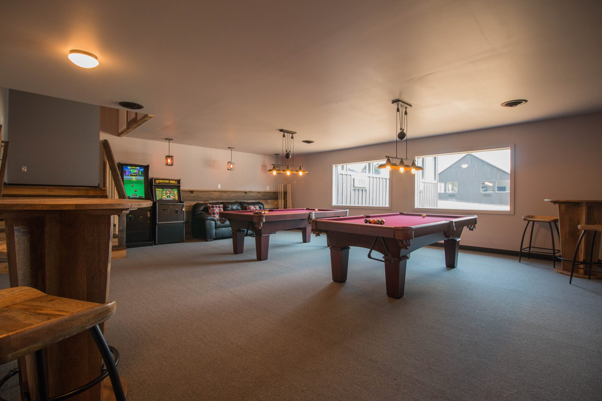 The billiard room: two pool tables, arcade cabinets and a bar rail with stools under a wide window