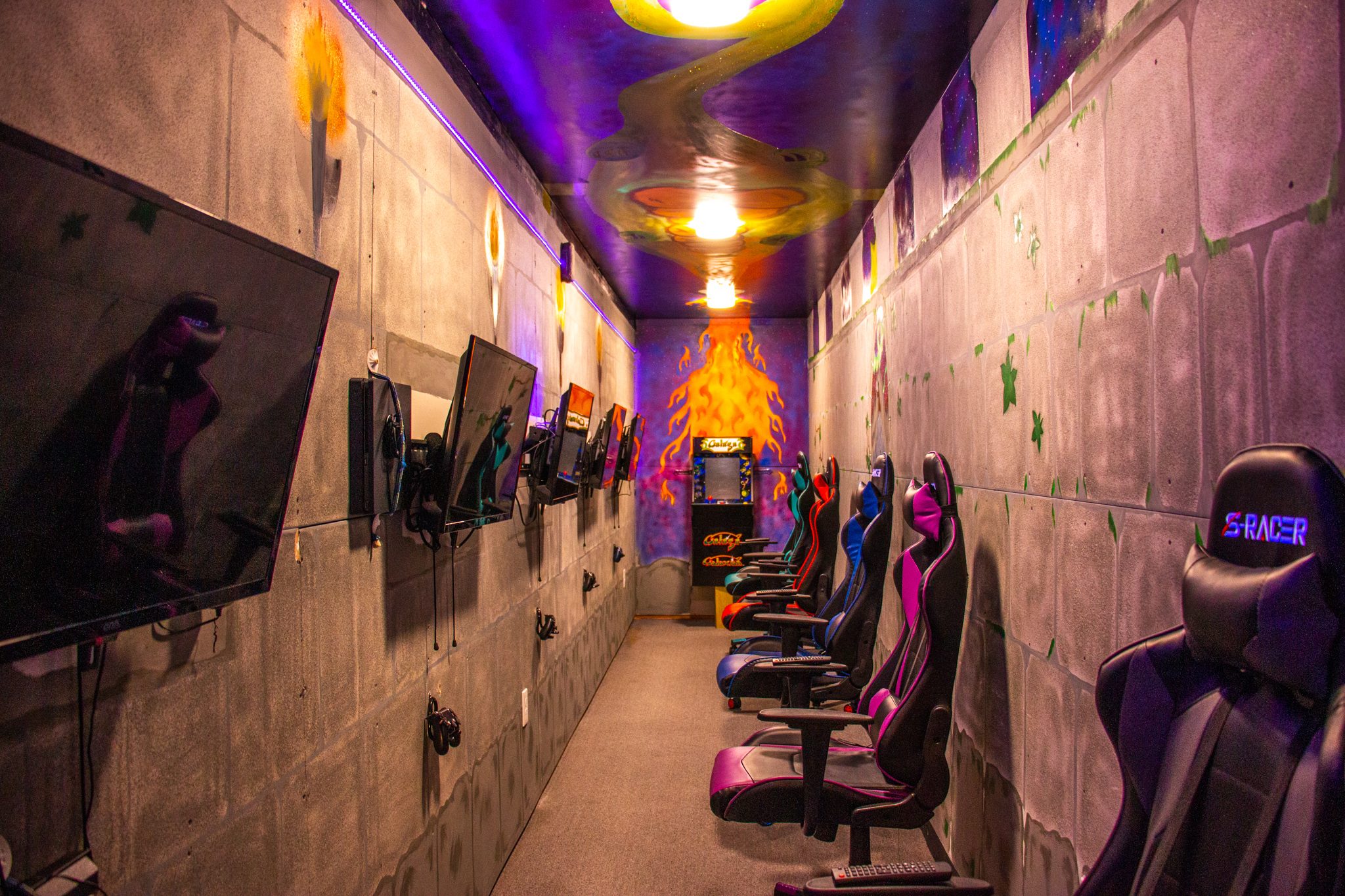 A long game room with gaming chairs facing a row of wall-mounted screens