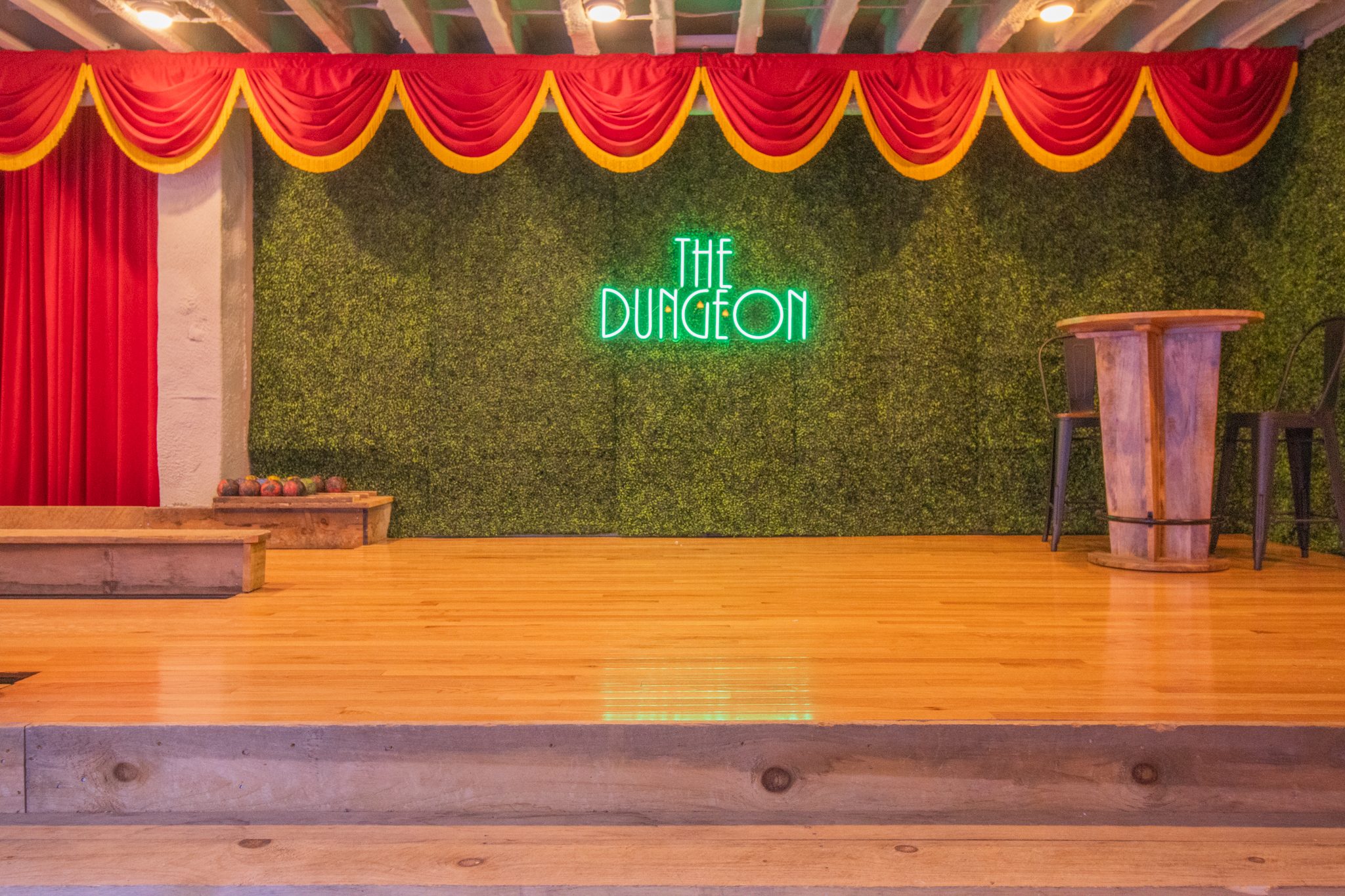 The stage under the theater screen: a raised wooden stage with red and gold curtains, a hedge wall and a neon sign reading The Dungeon