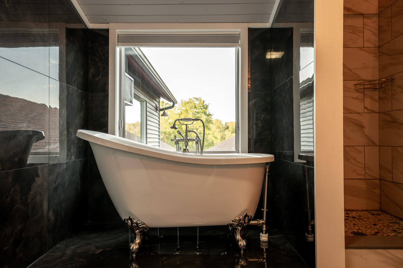 A clawfoot tub on a black-tiled floor, set against black tile below a tall window