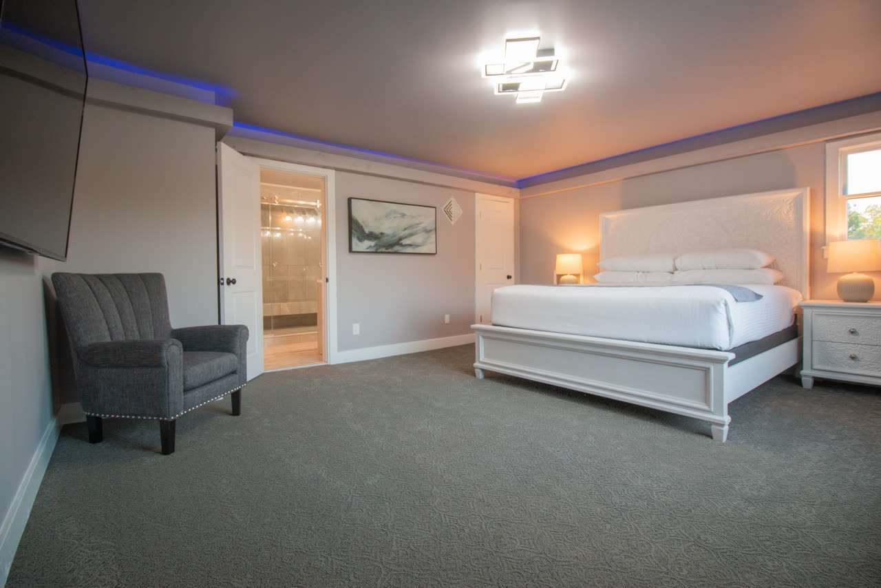 The master bedroom, a white king bed under a cool ceiling light, with the tiled bath through an open door