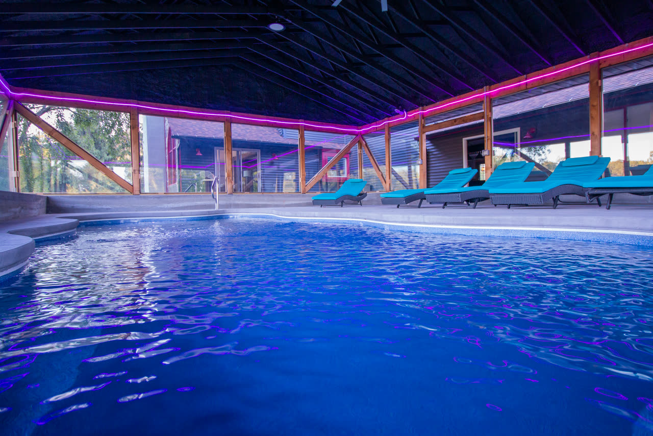 The indoor pool lit at night — blue water under a dark timber roof, magenta light running along the beams, and loungers on the far side