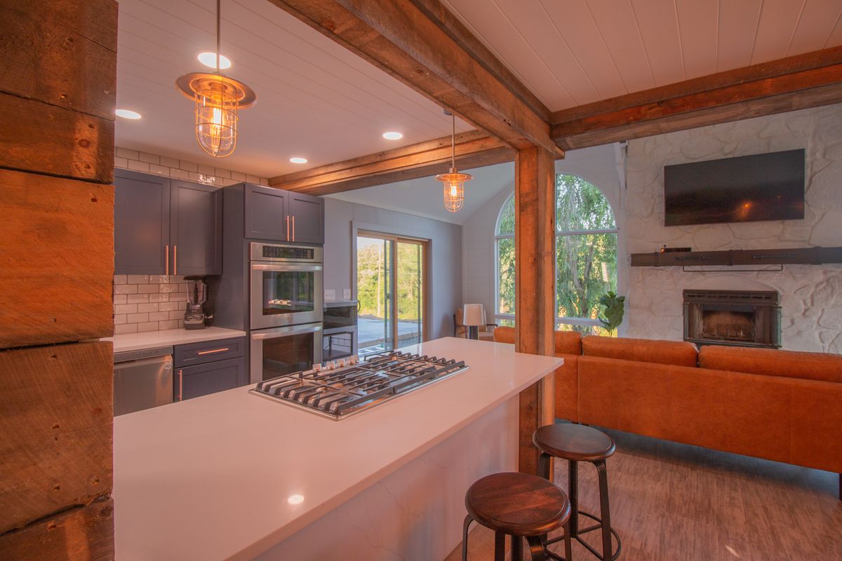 An open kitchen with a gas hob set in a white island and bar stools, a tan leather sofa beyond facing a white stone fireplace