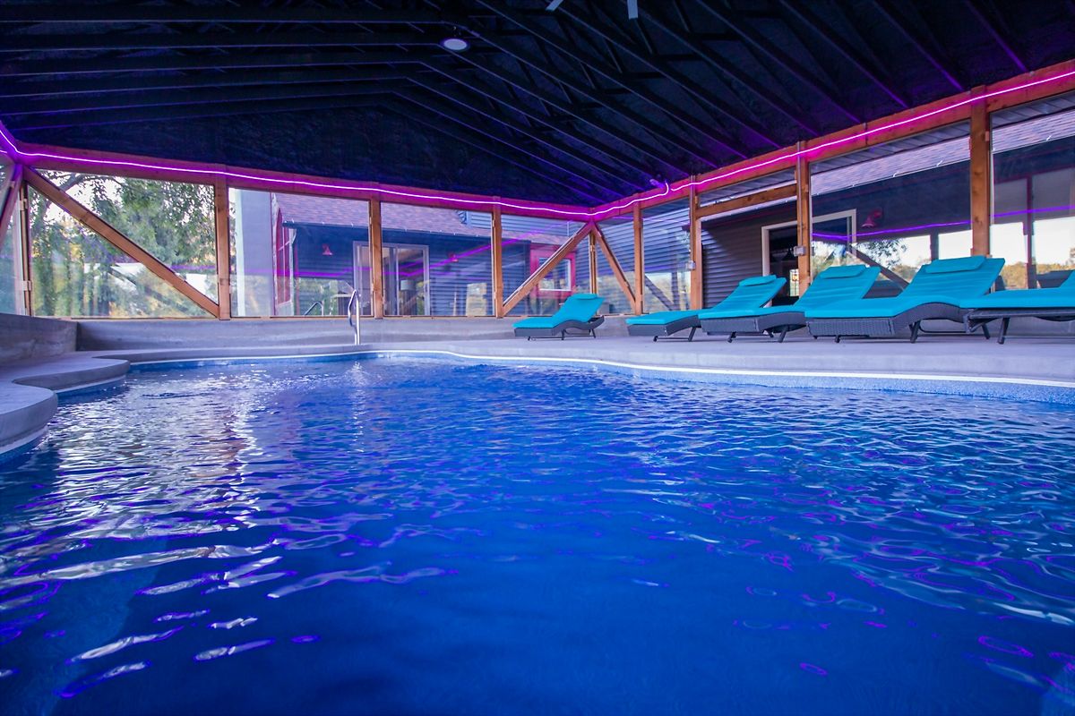 The indoor pool lit at night — blue water under a dark timber roof, magenta light running along the beams, and loungers on the far side