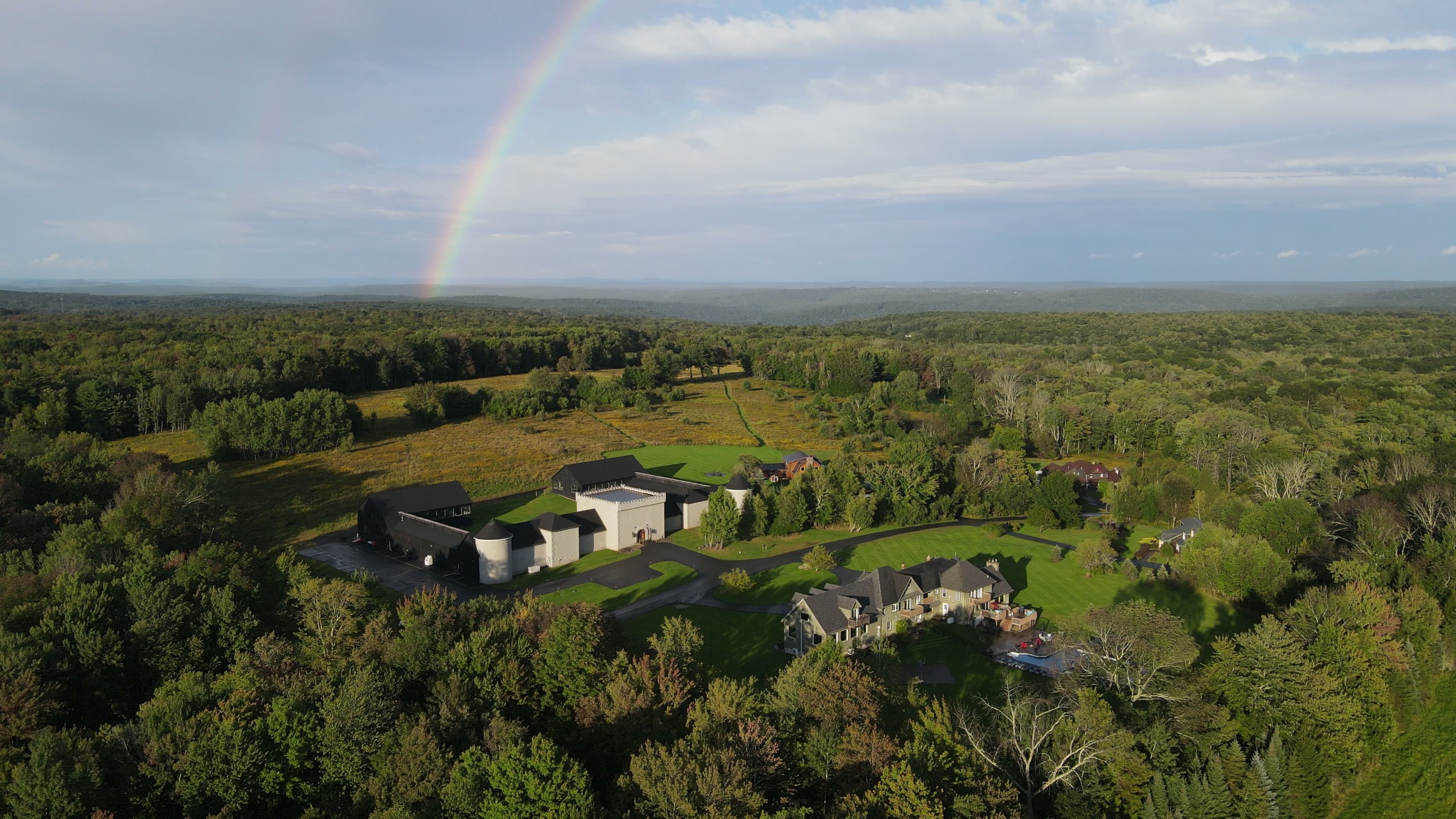 The Hilltop compound from the air in late-afternoon light, a rainbow coming down over the woods beyond the Castle and the Mansion