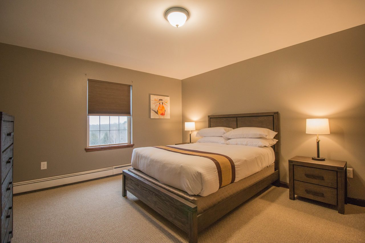 A bedroom with a queen bed between two lamps and a window with the blind half down