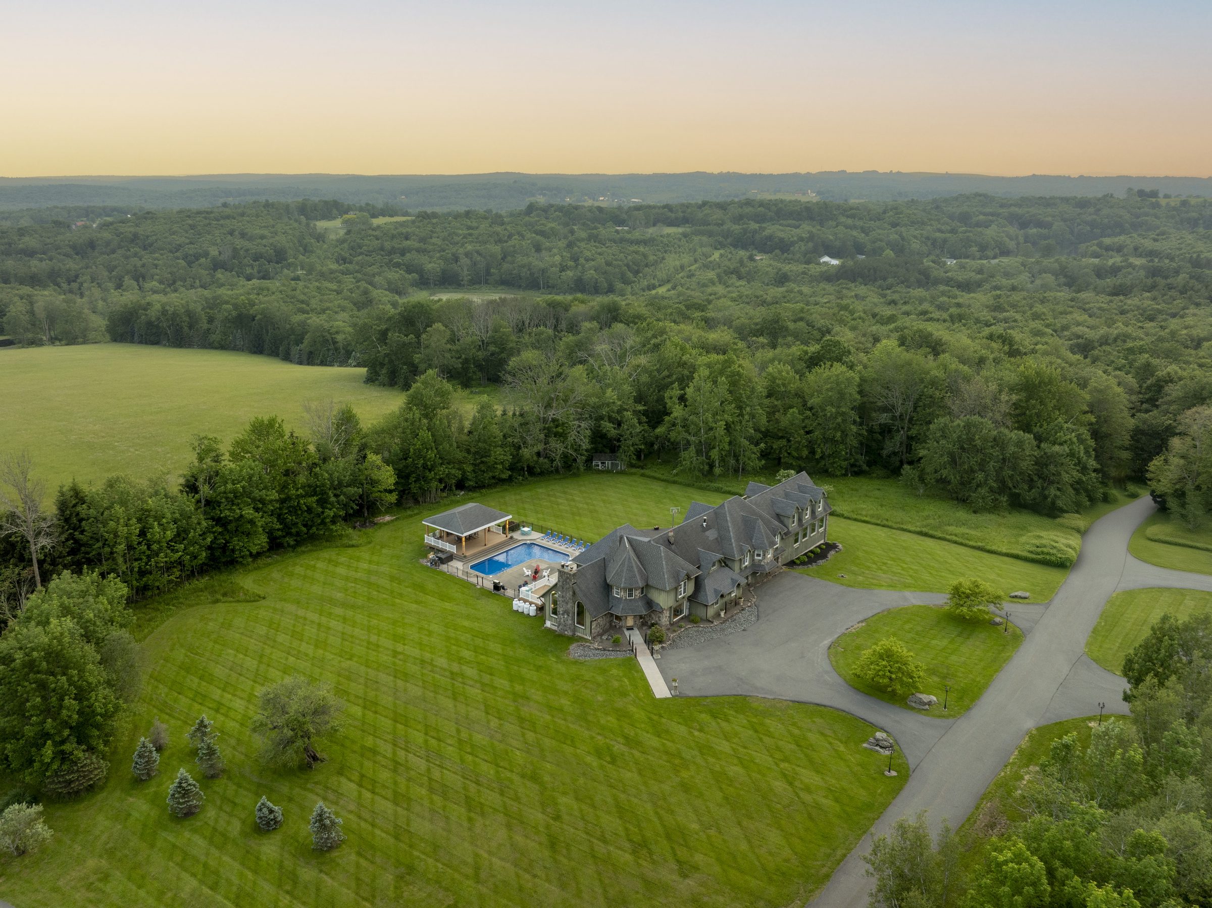 The Mansion from the air at dusk: the long gabled roof and its outdoor pool set in mown lawn, with the drive curving away and woodland beyond