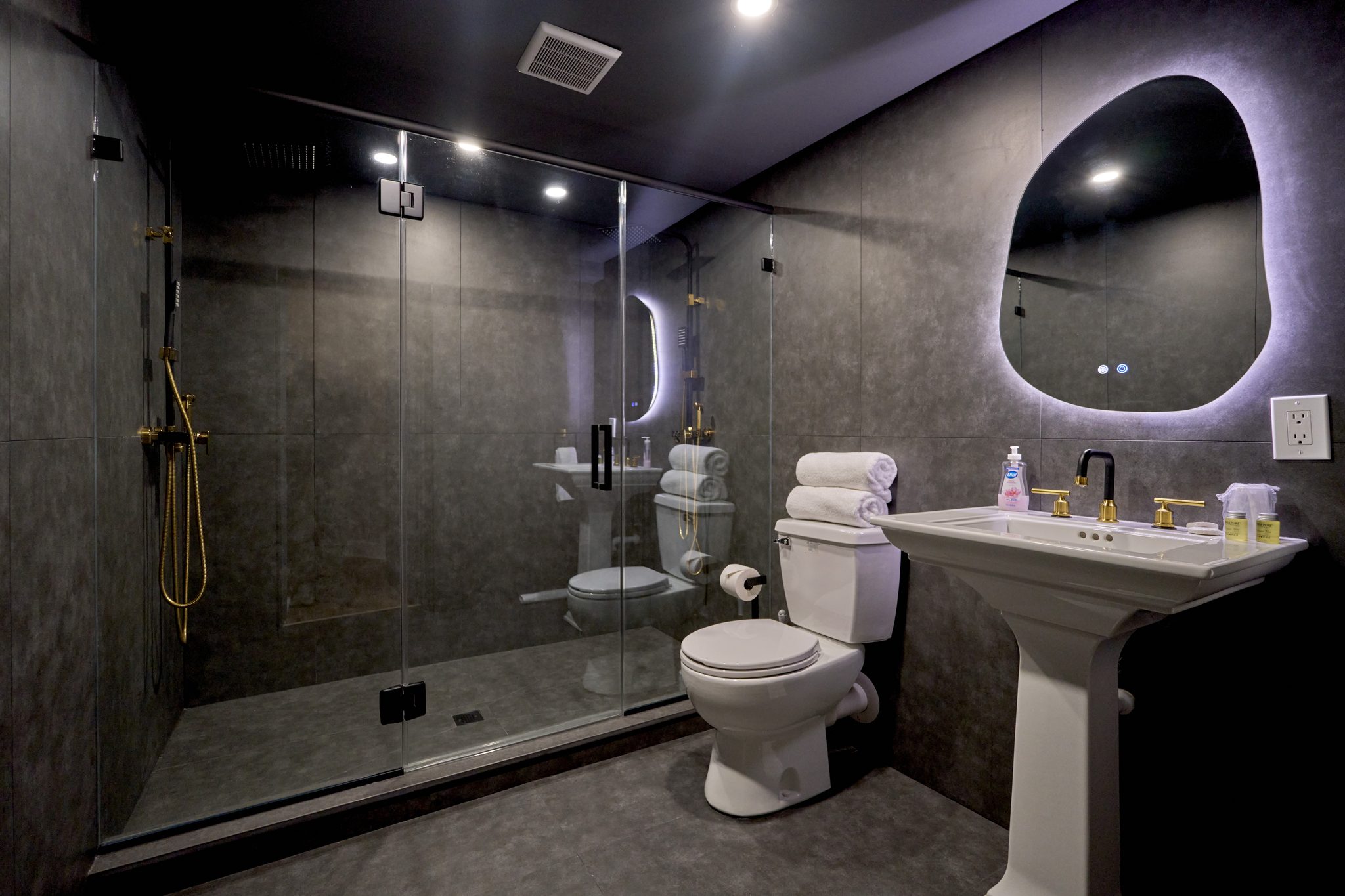 A dark-tiled basement bathroom with a walk-in shower, a pedestal basin and a backlit oval mirror