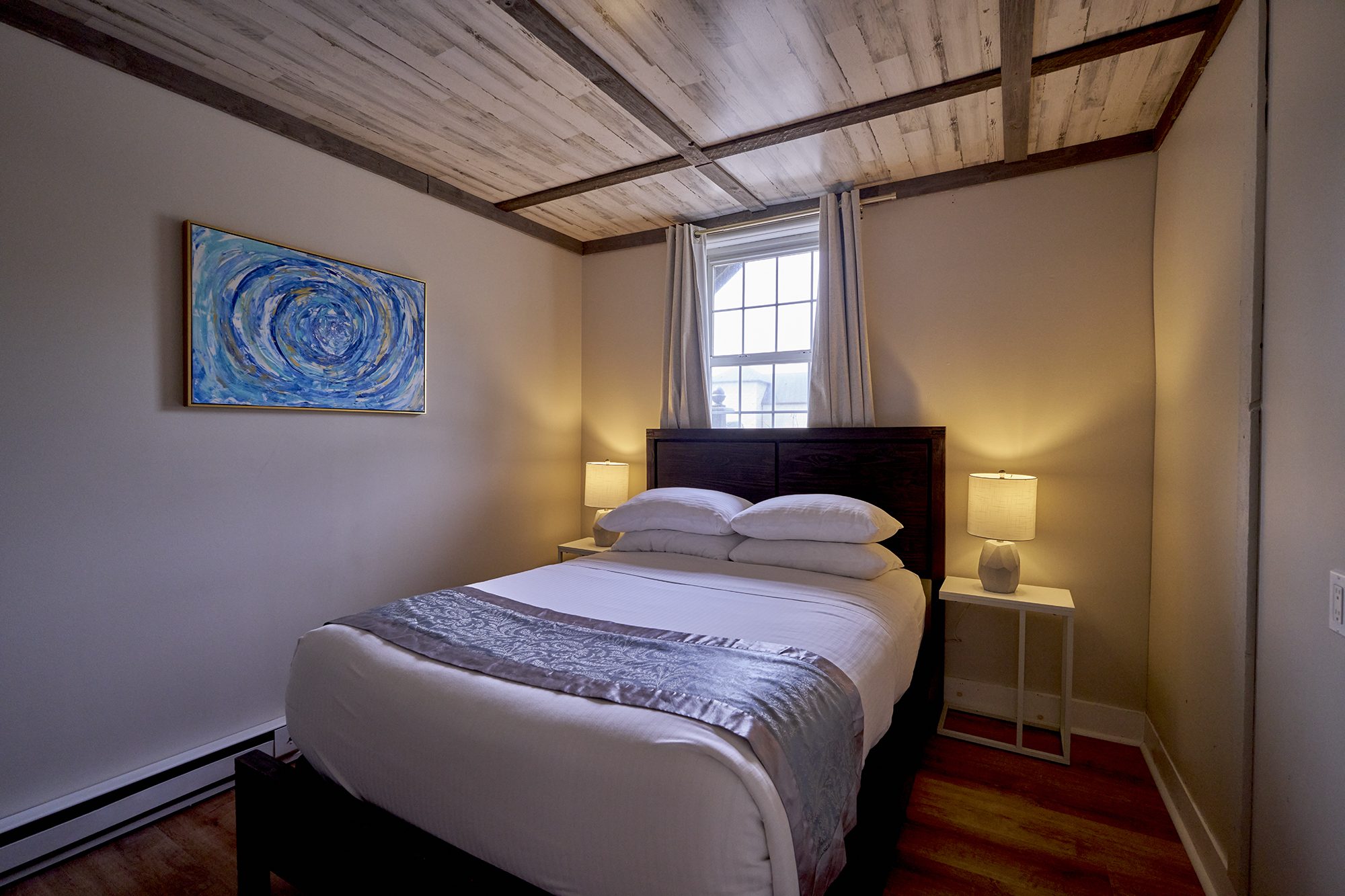 A queen bedroom with a plank ceiling and dark beams, a blue abstract over the bed