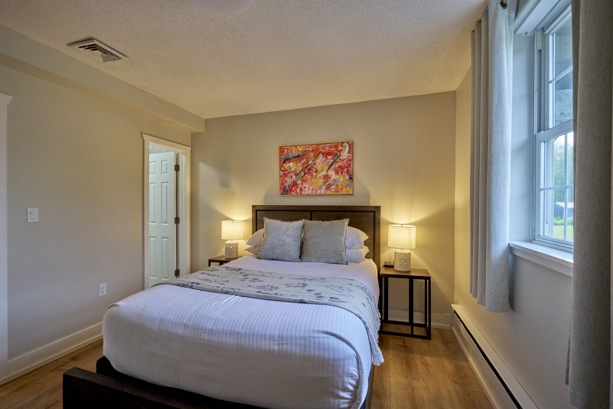 A queen bedroom with beige walls, white bedding and a window to one side, one of three near-identical rooms off the same hall