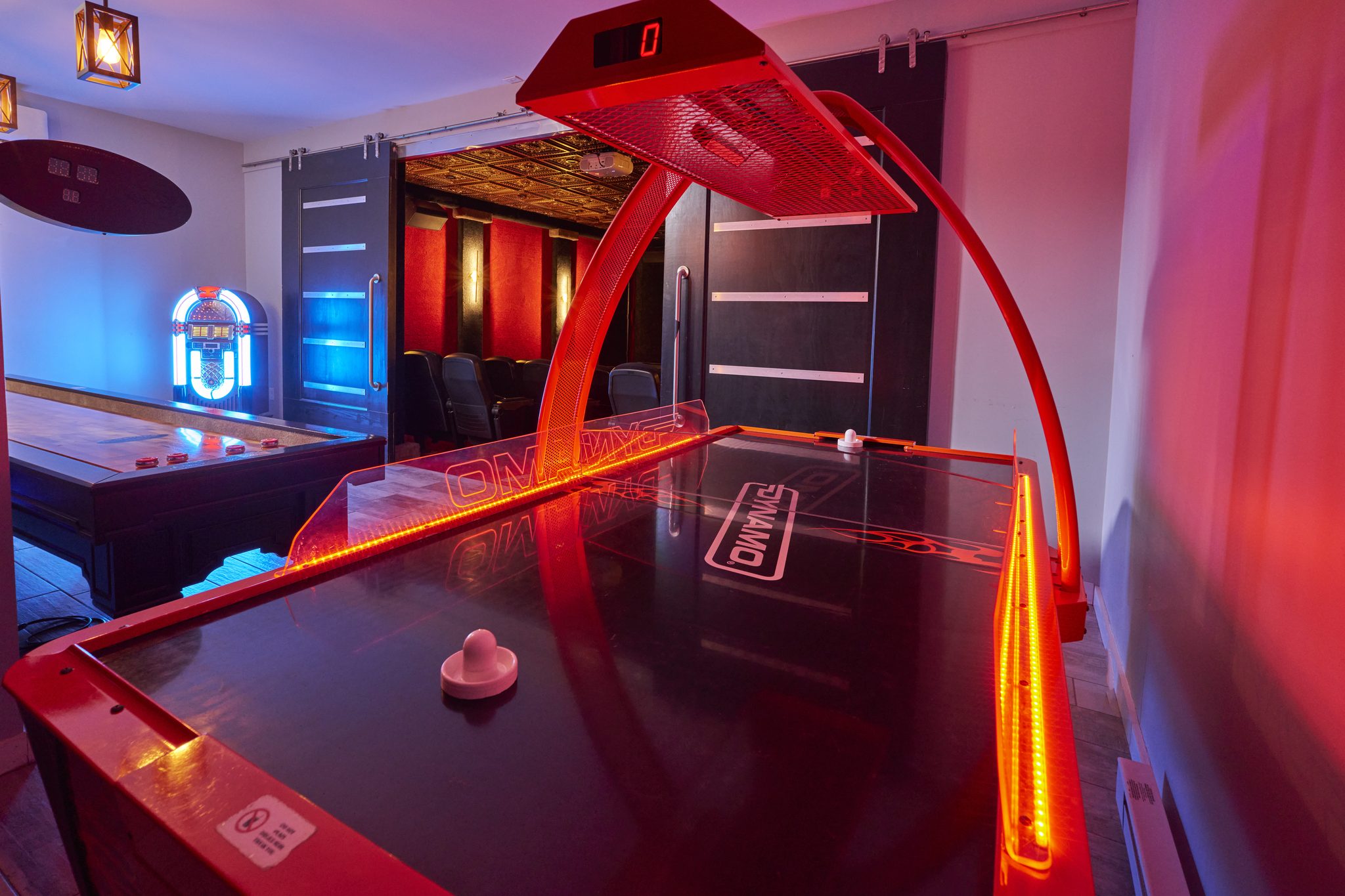 A game room lit red and blue, a shuffleboard table and a jukebox against a dark wall