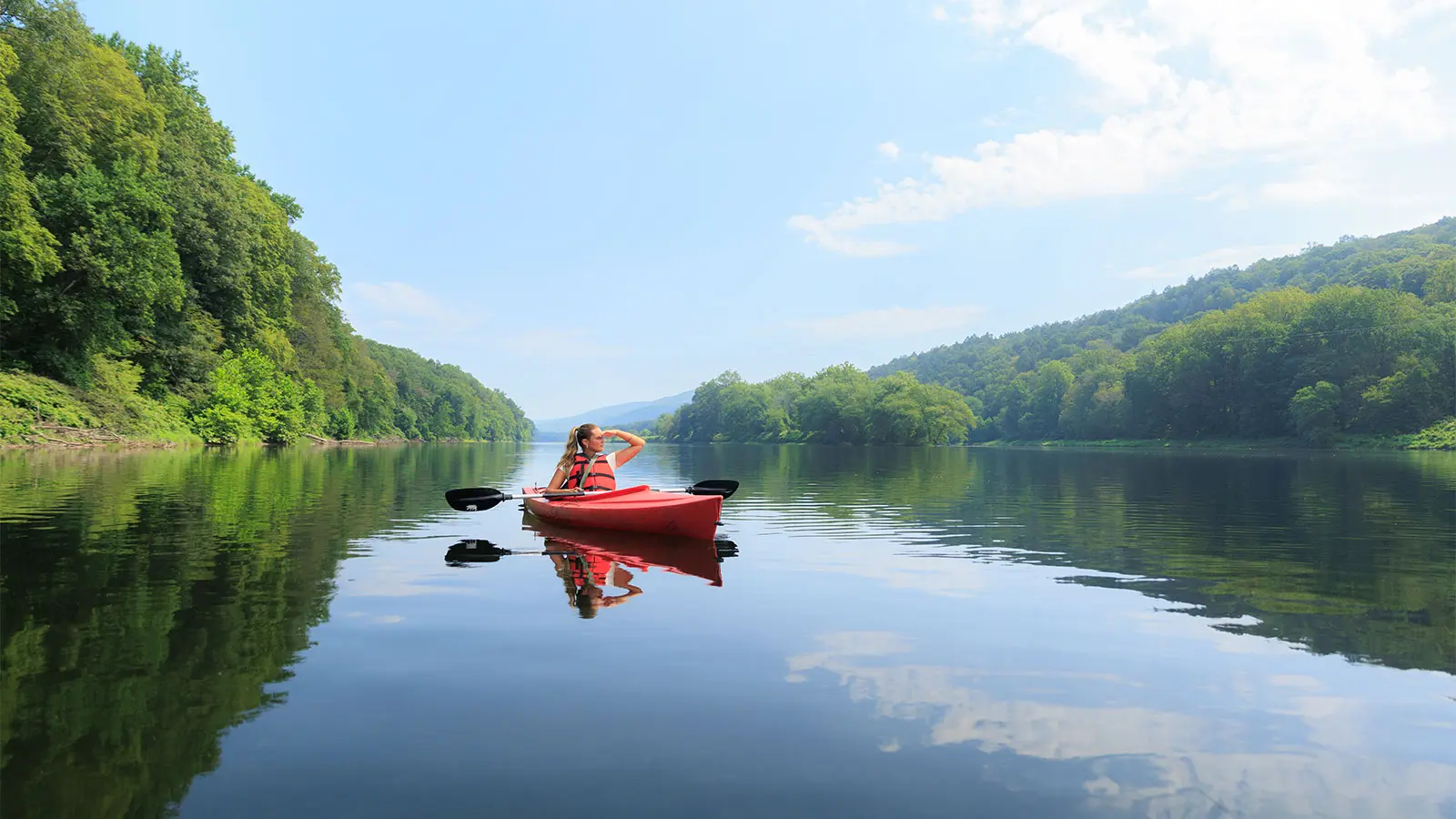 A woman paddling a red kayak alone down the middle of the Delaware, wooded banks either side and the sky mirrored in flat water